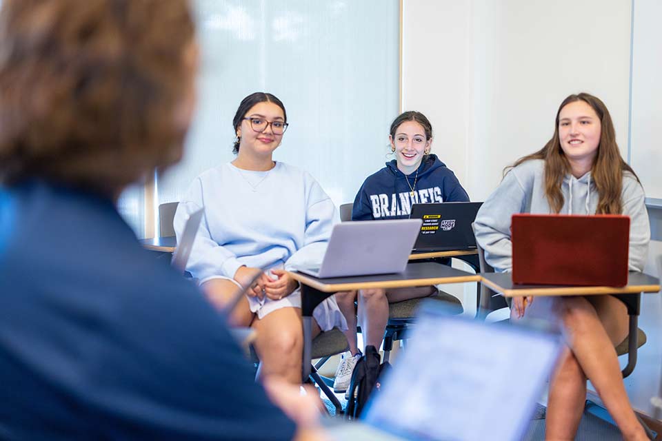 Three students seated at desks with laptops, listening to a professor speaking