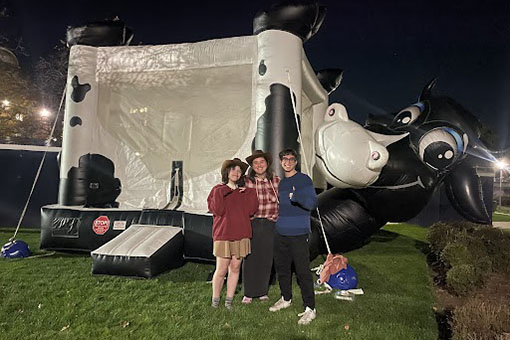 Three students posed in front of an inflatable bounce house shaped like a cow at a First Thursdays event.