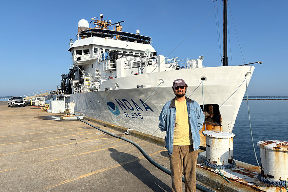 Greg Roitbourd '26 stands on a dock in front of a large NOAA research boat.