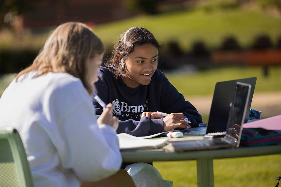 Two students sit outside working on laptops.