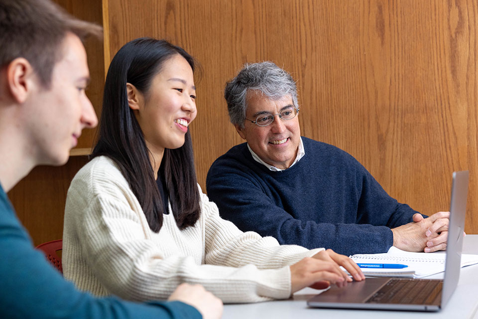A professor sits at a table and looks at a laptop with students