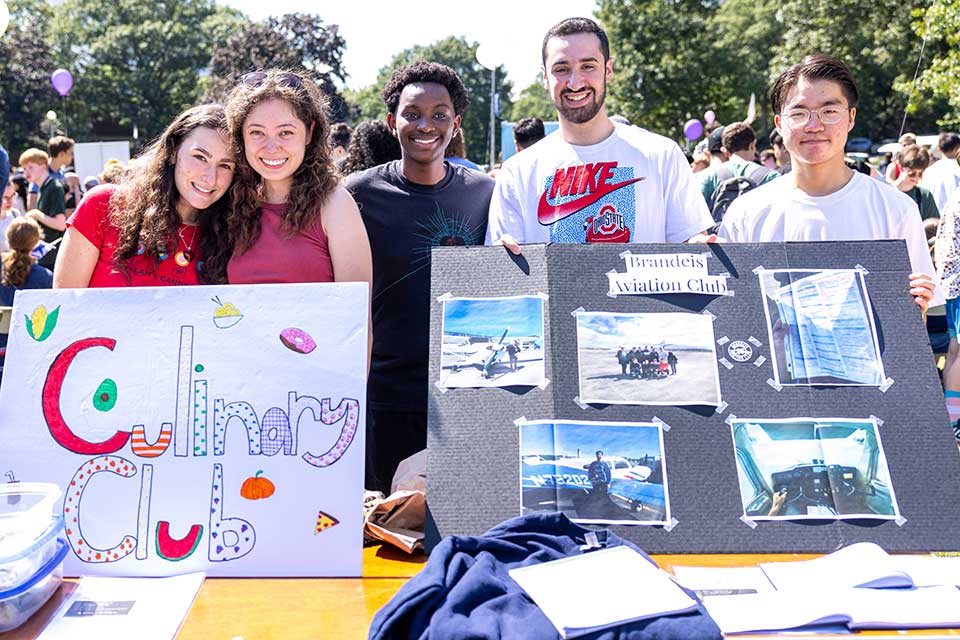 Five students smiling with two poster boards. One reads: Culinary Club. The other reads: Brandeis Aviation Club and contains pictures of people with aircrafts.