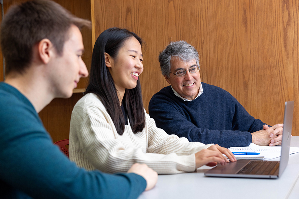 Two students and faculty member sit together, looking at a laptop and having a conversation.