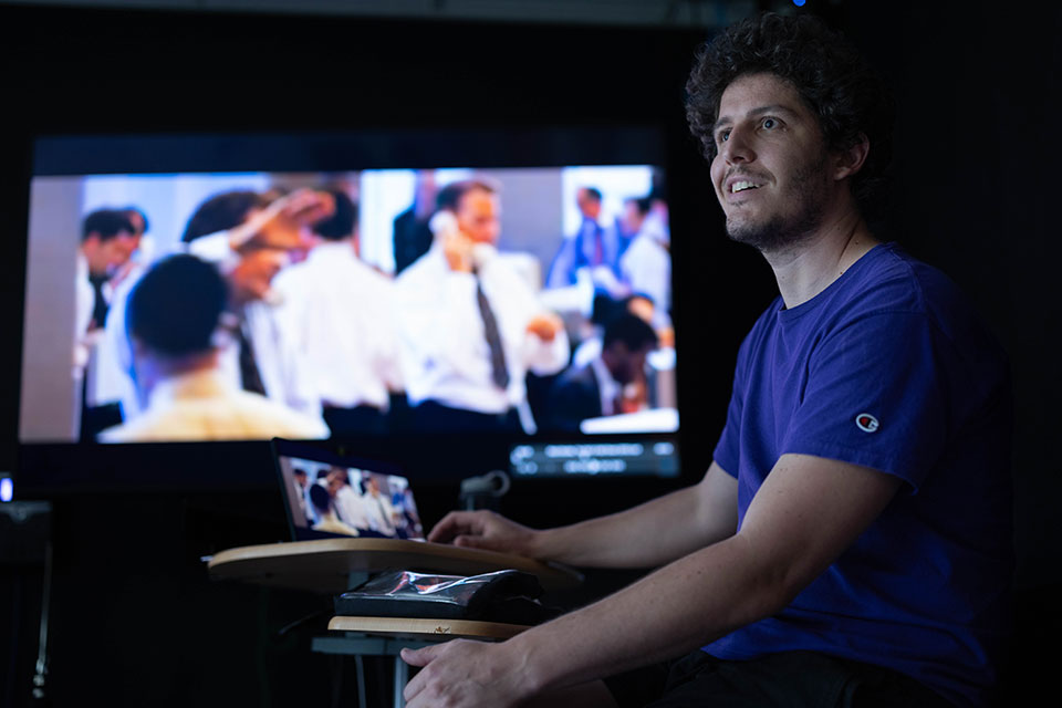 A film professor sits in a dark classroom with a screen playing a film in the background.