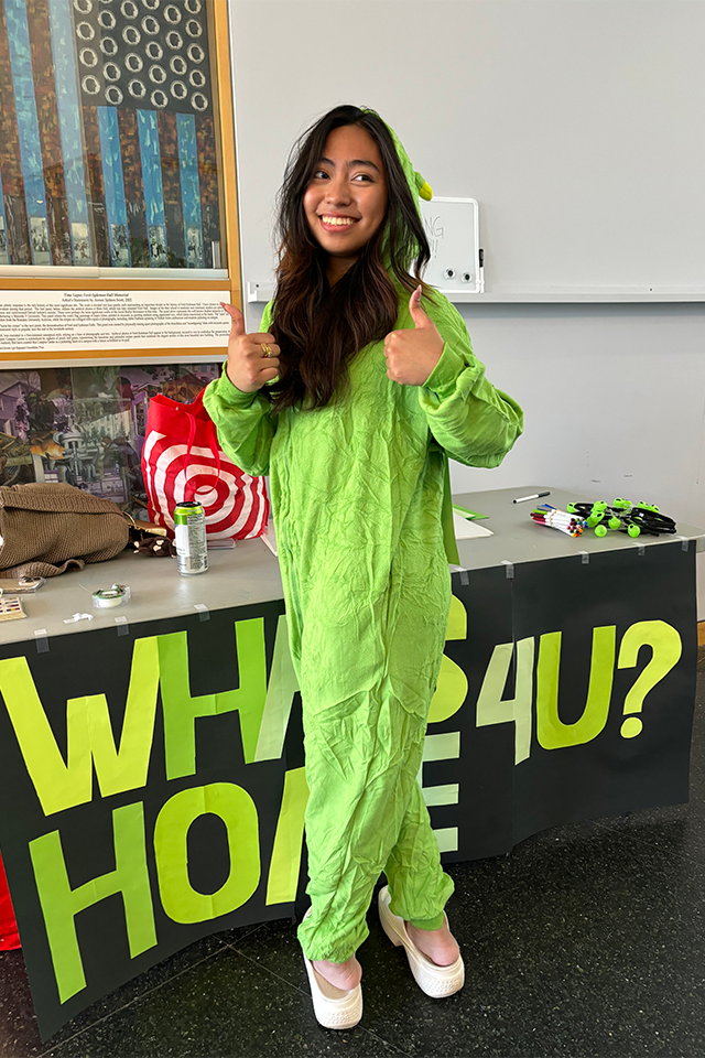 A young woman wearing a bright green “alien” costume stands indoors, smiling and giving two thumbs up. She is in front of a table with a large black sign with neon green letters reading “WHAT’S HOME 4U?” and various supplies on top.
